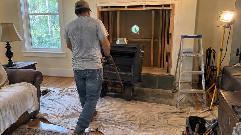 A worker wheels a new gas fireplace unit on a dolly toward a framed wall opening in a living room.