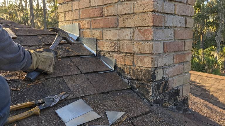 New step flashing pieces are slipped under brown shingles along the side of a tan brick chimney.
