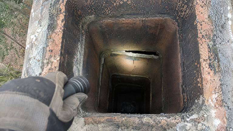 A flashlight beam down a sooty clay flue lights a cracked tile and an open joint below the top.