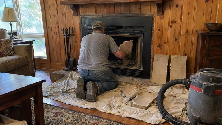 A worker lifts broken refractory panels out of a factory-built fireplace in a paneled den.