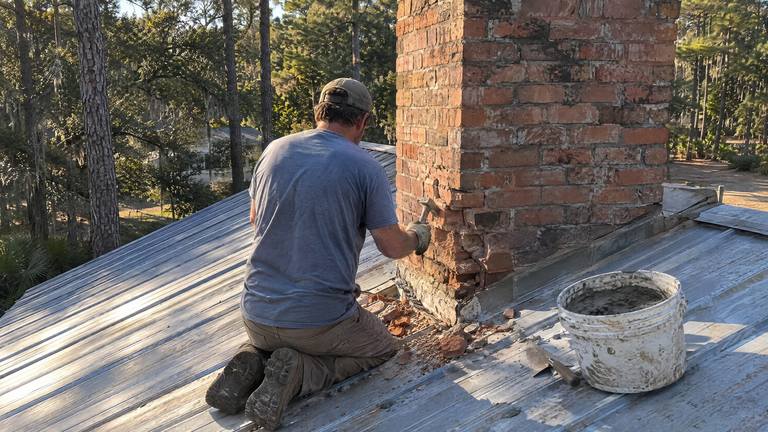 A worker kneels on a metal roof chipping crumbled brick from the lower corner of an orange-red chimney.