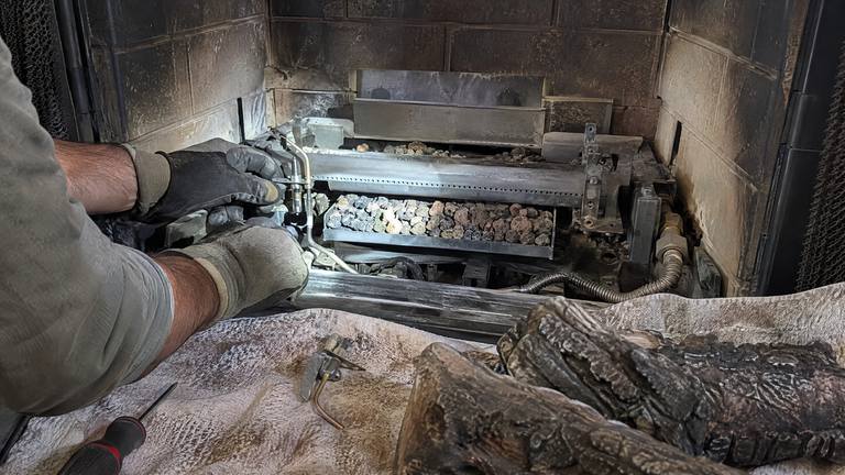Gloved fingers fit a new pilot assembly at the burner of a gas fireplace, with the logs lifted out.