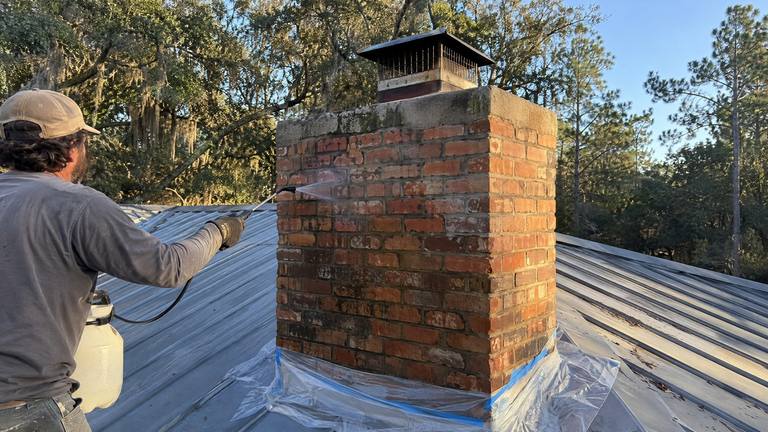 A worker sprays clear water repellent on an orange-red brick chimney with a pump sprayer on a metal roof.