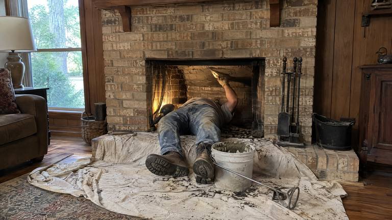 A worker lies on a drop cloth with his upper body inside a tan brick fireplace, reaching up into the throat with a trowel.