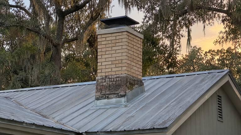 A tan brick chimney with a rebuilt top and a black cap above a metal roof, live oaks and an evening sky behind.