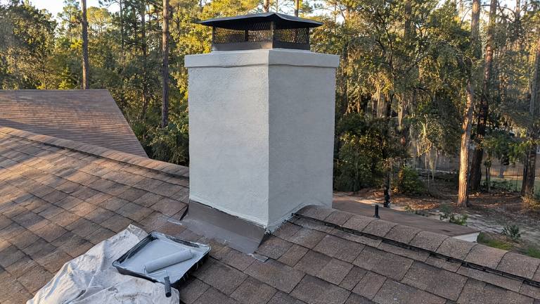 A stucco chimney with its cracks repaired and a fresh coat of pale gray paint above a shingle roof.