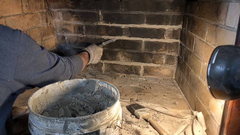 A gloved hand packs fresh mortar into eroded joints of a sooty firebox back wall.