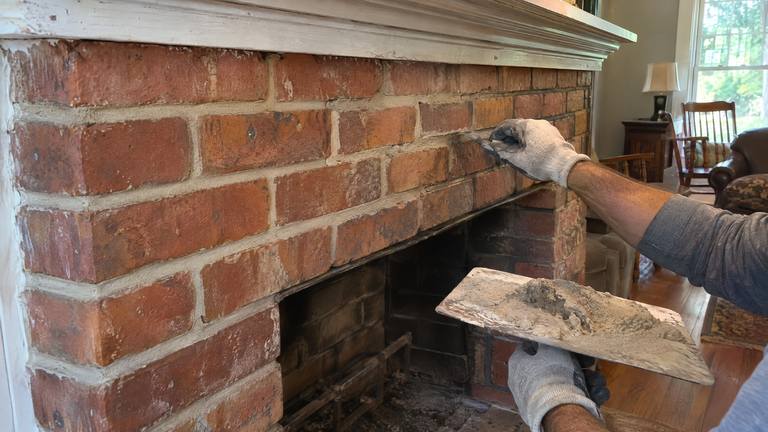 A gloved hand presses fresh mortar into the joints of an older orange-red brick fireplace face from a hawk.