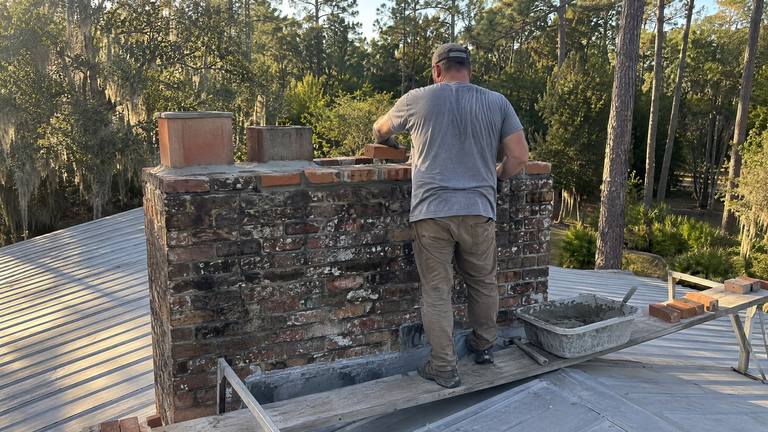 A worker on a metal roof resets the top bricks of an orange-red brick chimney with fresh mortar.