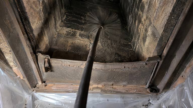 A round chimney brush on a rod works up through an open damper into a sooty flue, plastic sheeting taped around the opening.