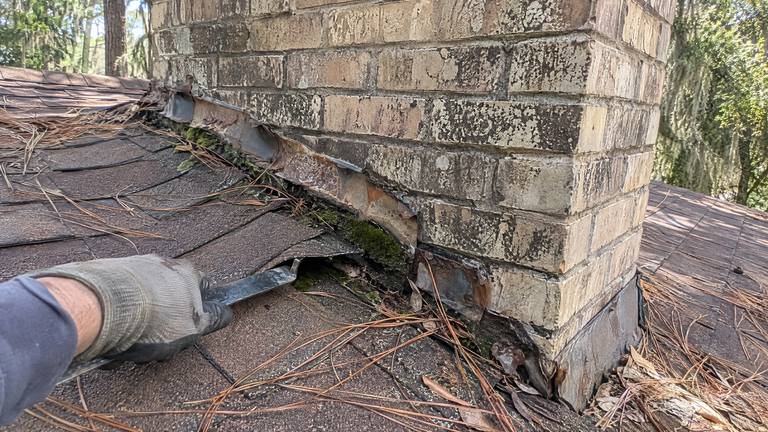 Soft, rotted shingles and rusted flashing at the base of a tan brick chimney on a shingle roof.