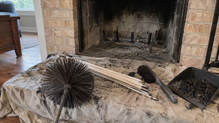 A sooty round chimney brush and a bundle of rods lie on a drop cloth in front of a tan brick fireplace.