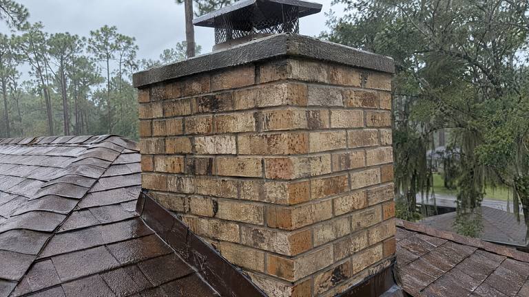 A tan brick chimney with a black cap above wet brown shingles on a gray, rainy day.