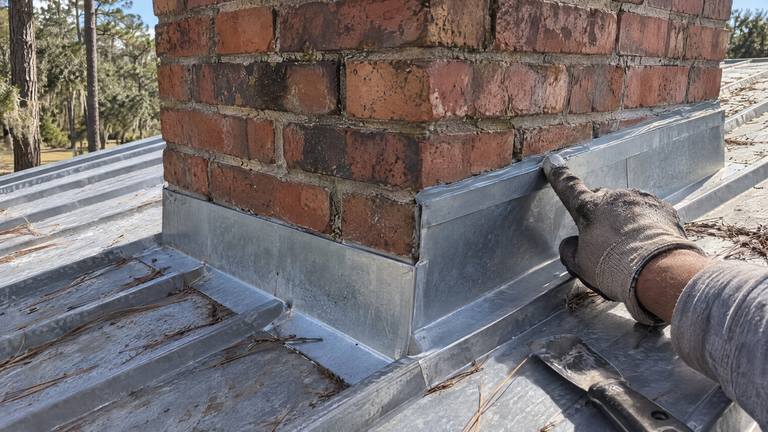 Gloved hands press sealant along new bent metal flashing where a brick chimney meets a galvanized metal roof.