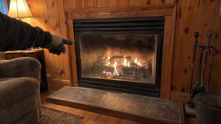 A gloved hand points at soot darkening the glass of a lit gas fireplace in a pine-paneled den.