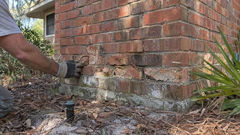 Crumbling, flaking bricks near the ground at the base of an exterior orange-red brick chimney.