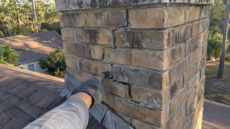 A stair-step crack and shifted bricks in the mortar joints of a weathered tan brick chimney, with a gloved hand at an open joint.
