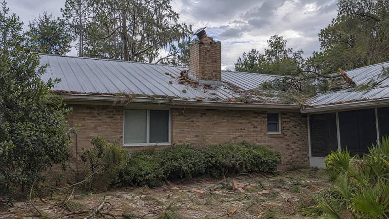 Loose bricks from a broken chimney top and fallen pine branches lie across a metal roof after a storm.