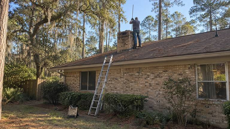 A worker on a brown shingle roof pushes a sweep rod down into a tan brick chimney, a ladder against the eave below.