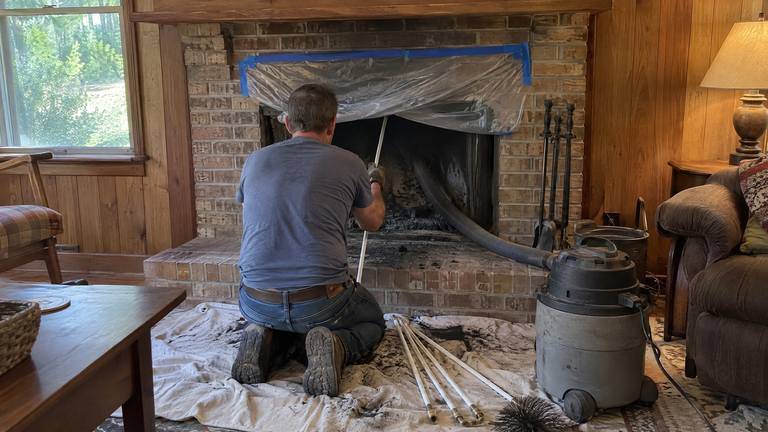 A worker kneels at a tan brick fireplace feeding flexible sweep rods up the flue, with a vacuum hose at the opening.
