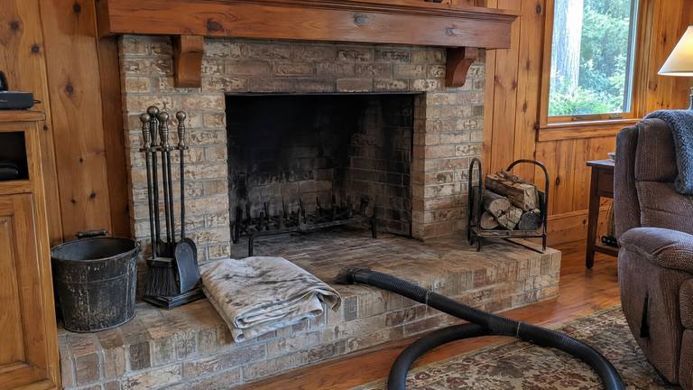 A swept tan brick fireplace in a pine-paneled den with a folded drop cloth and a vacuum hose on the hearth.