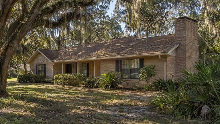 A tan brick ranch home with a wide end chimney in the shade of large live oaks with Spanish moss.