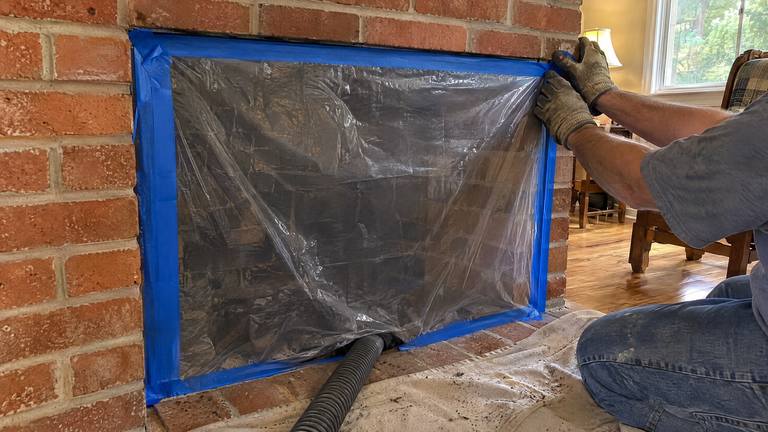 Gloved hands tape plastic sheeting over a fireplace opening before a chimney sweep.
