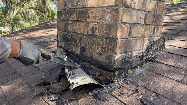 Old tar-smeared flashing is peeled away from the base of a brick chimney on a shingle roof.