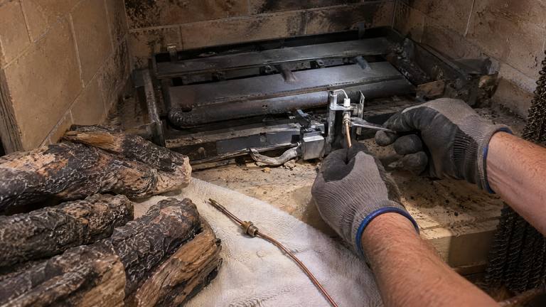 Gloved hands tighten a fitting at the pilot of a gas log burner, the ceramic logs lifted out onto a towel.