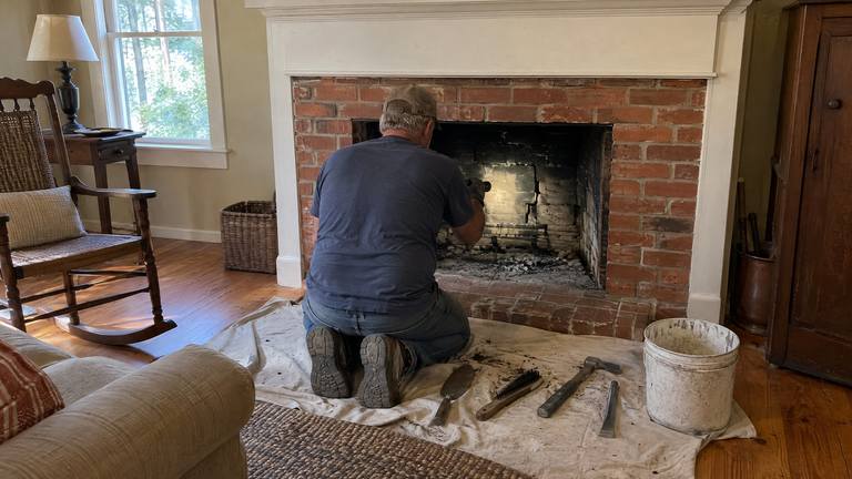 A worker kneels at an orange-red brick fireplace with repair tools laid out on a drop cloth.