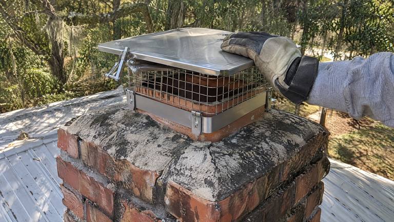 A gloved hand closes the lid of a stainless top-sealing damper mounted on a clay flue.