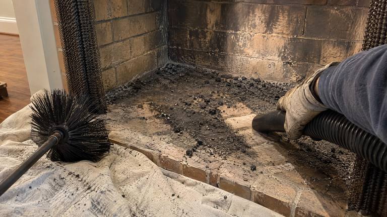 A gloved hand vacuums loose soot from the floor of a tan brick firebox after a chimney sweep.