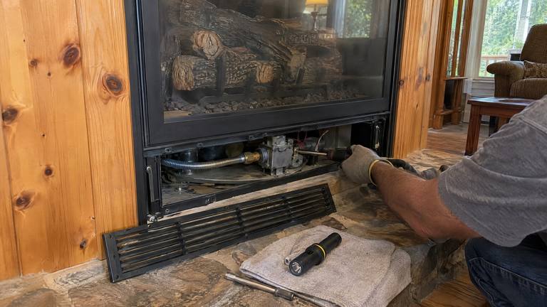 The lower louver panel is off a gas fireplace, showing the valve, while a gloved hand holds a nut driver.