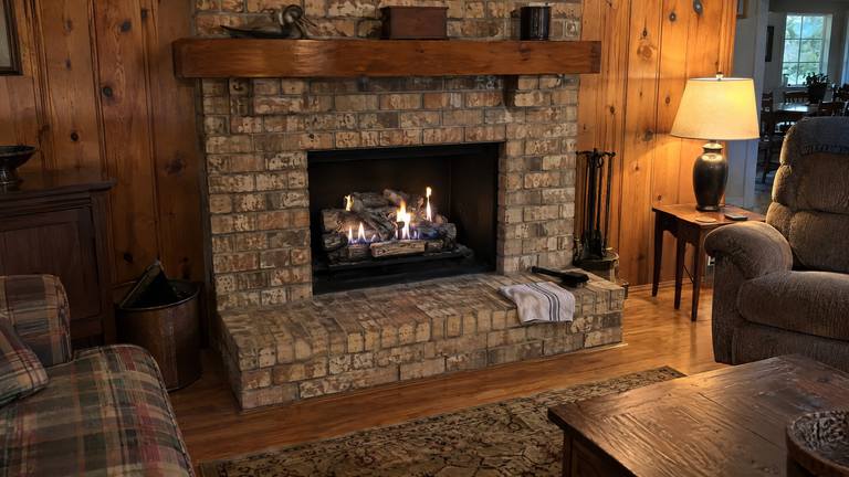 A vent-free gas log set burns with short blue and yellow flames in a tan brick fireplace in a paneled den.
