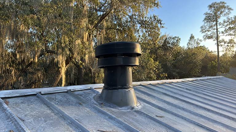 A round black gas fireplace vent cap rises through a metal roof on a short pipe with a flashing boot.