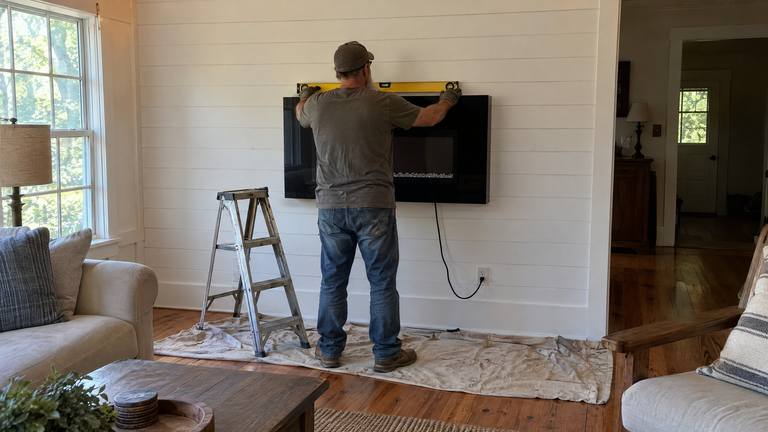A worker holds a level on a wall-mounted electric fireplace newly hung on a painted board wall.