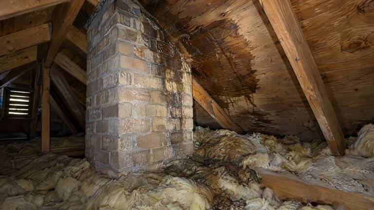 Dark wet stains on attic roof boards beside a tan brick chimney, lit by a flashlight.