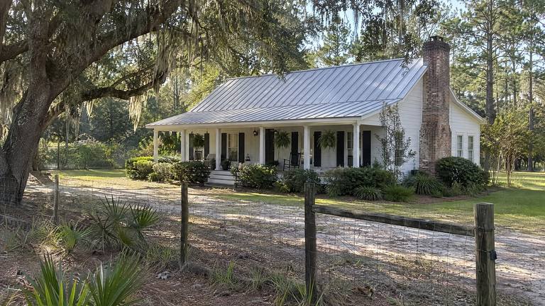 A white frame farmhouse with a deep front porch, a metal roof and a tall brick chimney on the gable end behind a wire fence.