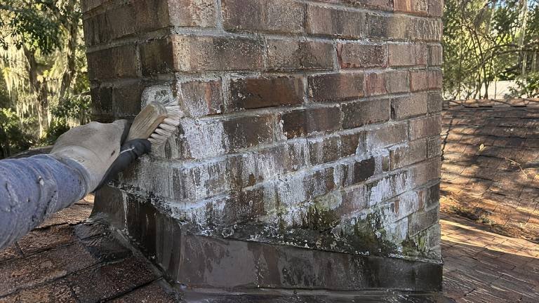 White mineral bloom streaks the lower courses of a brick chimney while a gloved hand brushes it with a stiff brush.