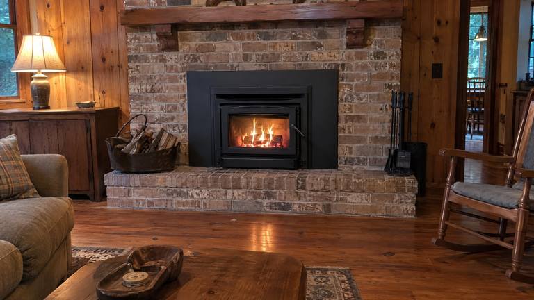A black wood-burning insert with a fire behind its glass sits in a tan brick fireplace in a paneled room.